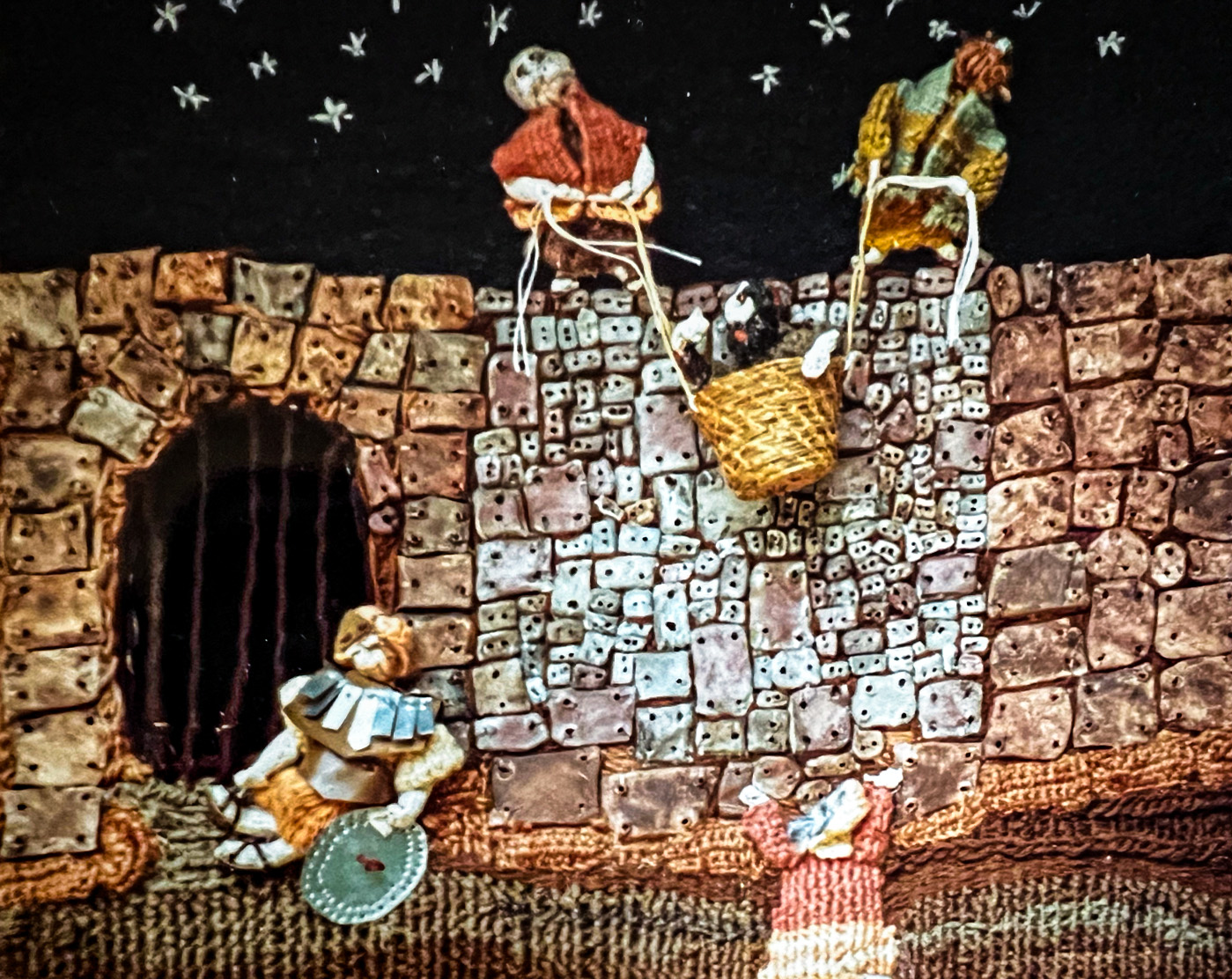 Fiber art depicting Paul being lowered in a basket over a stone wall at night.